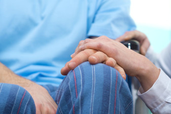 En bild på två personers händer. Den ena personen sitter i rullstol och den andra personens hand vilar på den rullstolsburnes hand.