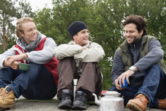 Tre män som verkar vara vänner sitter på en klippa i naturen. De ser glada ut.