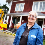 Kvinna står i målarkläder framför en hembygdsgård.